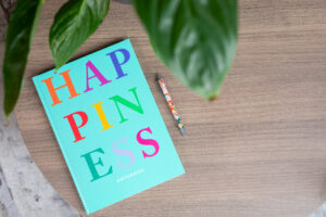 A large blue/green notebook laying on a desk. The notebook has the word HAPPINESS in large letters on the front.